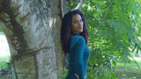 an east indian model with curly hair stands next to a large tree trunk in green dress