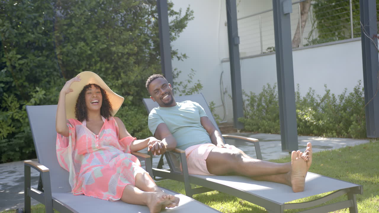 Relaxing outdoors, african american couple holding hands and lounging on poolside chairs