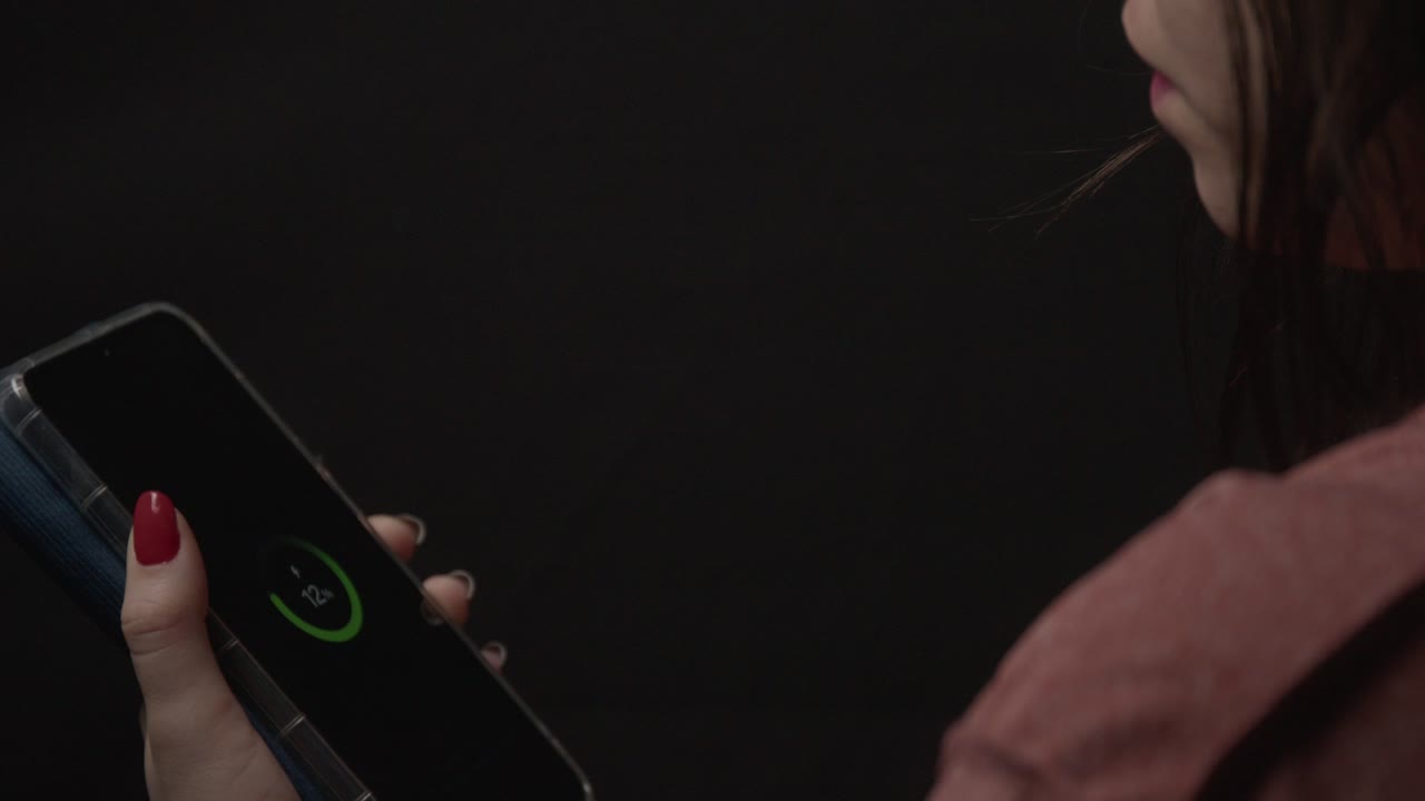 A woman holds a smartphone showing a charging symbol in a dimly lit room, highlighting red nail polish and casual attire. Intimate ambiance with focus on technology