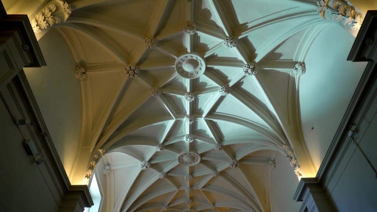 Telecamera inclinata che si muove verso l'alto attraverso una volta storica, rivelando archi a crociera, chiavi di volta e un cleristorio.