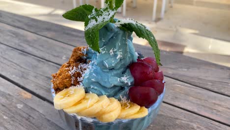 delicious blue spirulina frozen fruit bowl with bananas, homemade cookie crumble, grapes and mint on a wooden table, 4k shot