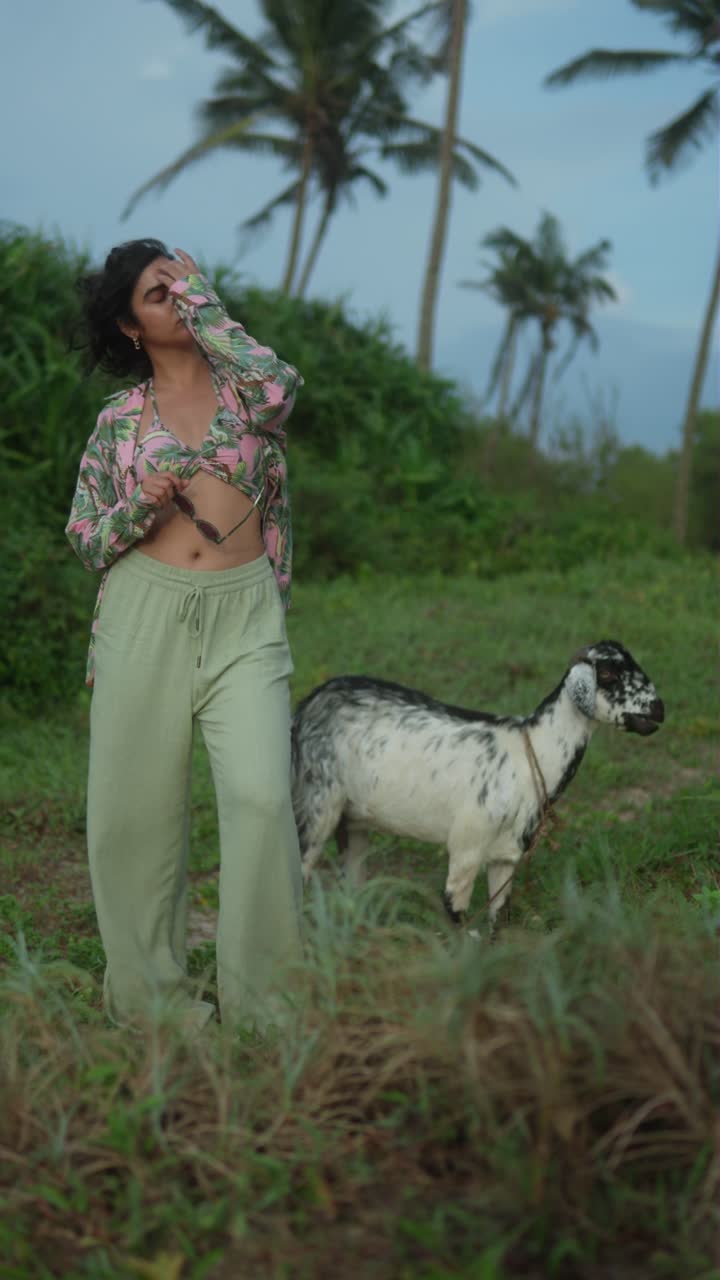 Woman in tropical outfit with a goat nearby in a lush outdoor setting, vertical shot
