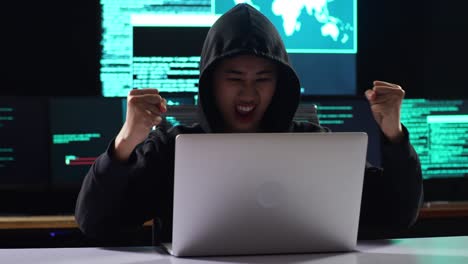 hacker asiática con capucha celebrando el éxito del ataque a los servidores de infraestructura global por un portátil con mapas y datos en pantallas de múltiples monitores en una habitación de oficina oscura