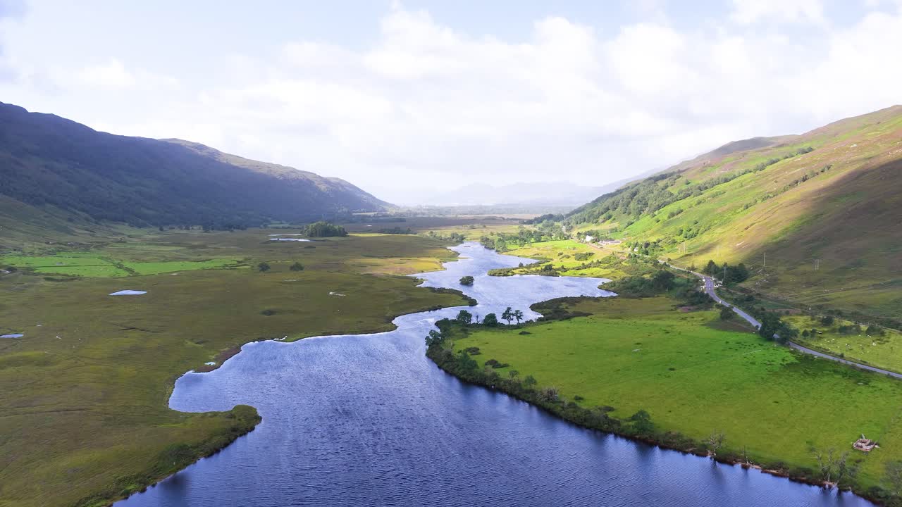 Drone glides over tranquil river valley, lush meadows, and mountains under soft daylight