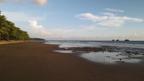 Dron,-Vuelo-Bajo-Medio,-Playa-De-Arena,-Olas-Pacíficas,-árboles-Verdes,-Cielo-Nublado-En-Calma