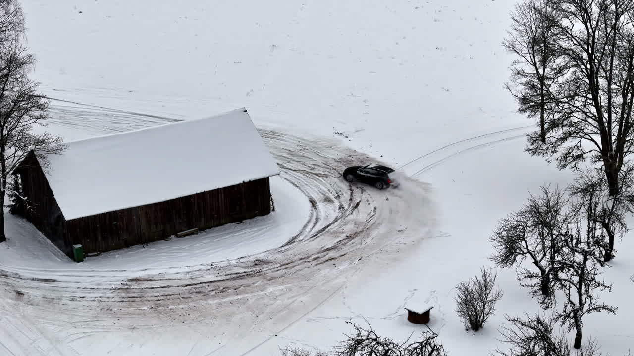 Aerial view of a car sliding on a snowy path beside a dark barn, leaving tire tracks in a vast white winter landscape