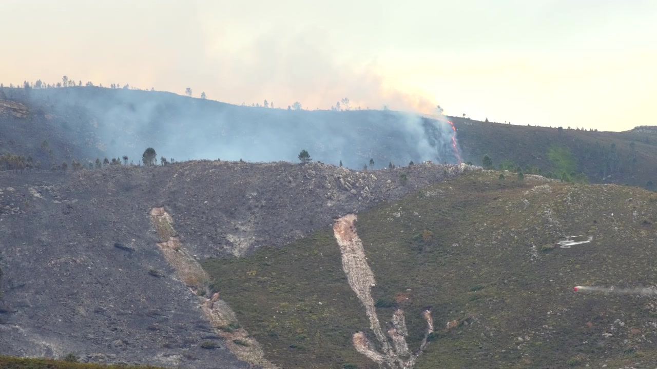 Incendio boschivo fuori controllo sul fianco della montagna mentre un elicottero con benna antincendio passa.