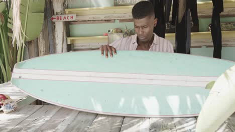 african american man preparing surfboard on the counter of surf rental beach shack, slow motion