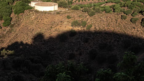 Aerial-view-of-a-dry,-hilly-landscape-with-scattered-greenery-and-a-water-tank,-shadow-of-clouds-passing