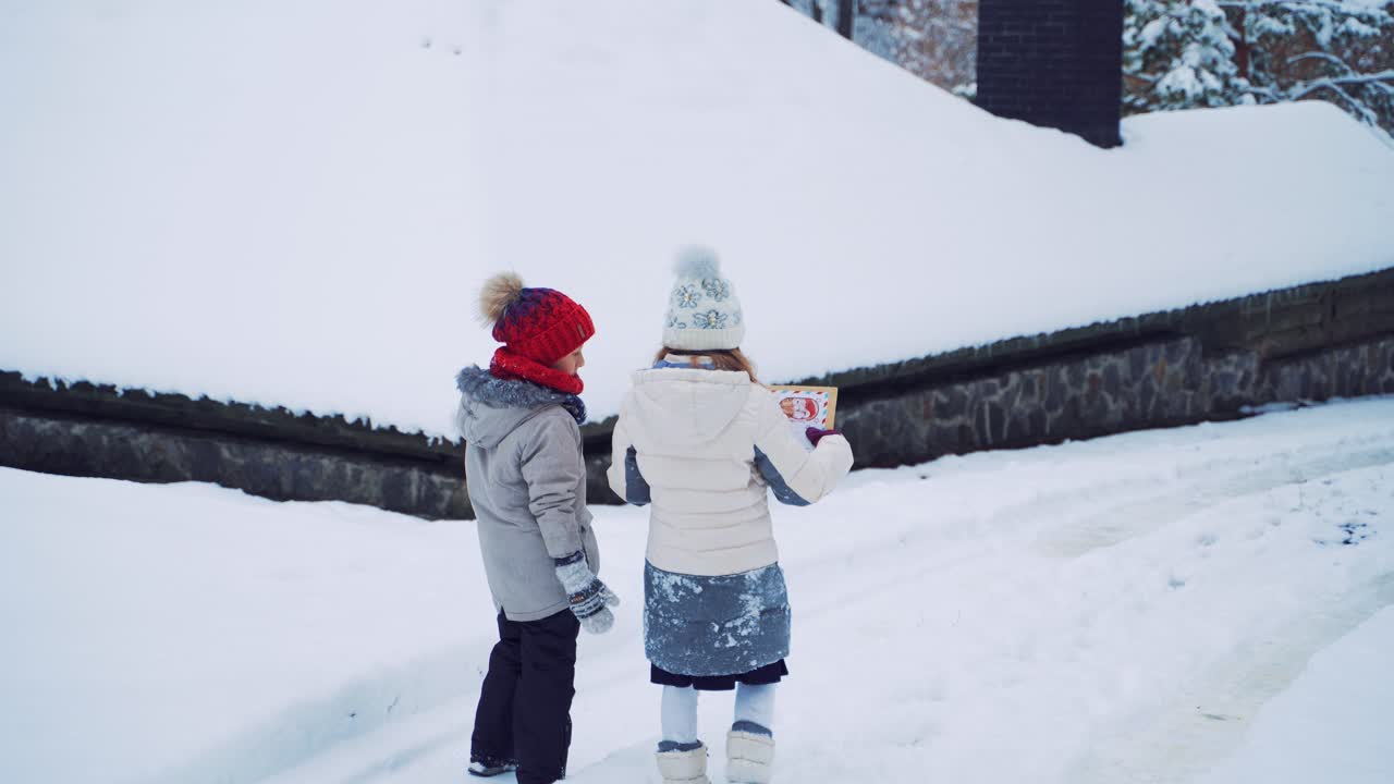 Children stop near the snowy house and talk in winter. Back view of two little kids walking in beautiful white snow outdoors. Children in warm clothes look into Christmas card.