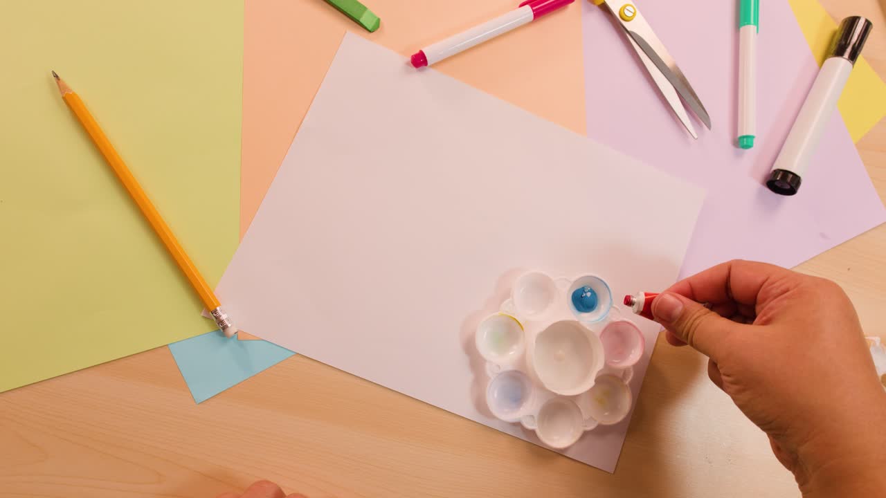 Hand places blue, red, yellow paint into palette on art desk with soft overhead lighting