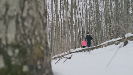 Positive-beautiful-young-healthy-couple-running-with-sportswear-through-the-forest-in-the-sunny-winter-morning.-Jump-over-the-tree,-overcome-the-difficulties-of-the-path.-step-over-an-obstacle