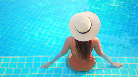 back view of unrecognizable woman in orange monokini and sunhat sitting on the border of hotel swimming pool inside water, slowmotion elevated point of view static