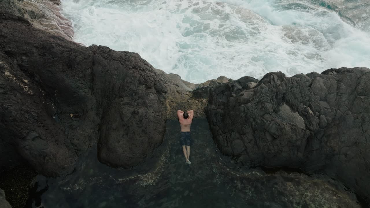 Top-down aerial of a man floating peacefully in volcanic tide pools near crashing ocean waves in Seixal, Madeira.