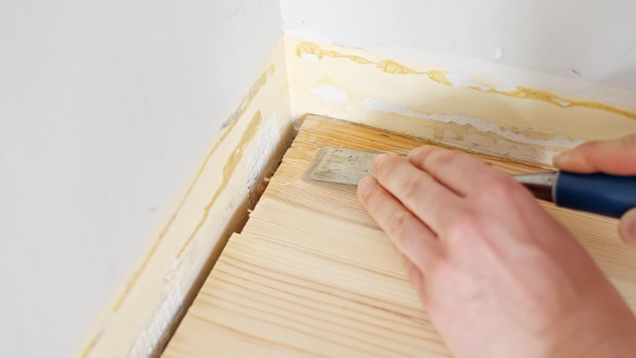 Close-up footage of detailed manual floor restoration work showing careful scraping of wooden flooring corner to remove old adhesive residue using a metal scraping tool in renovation context