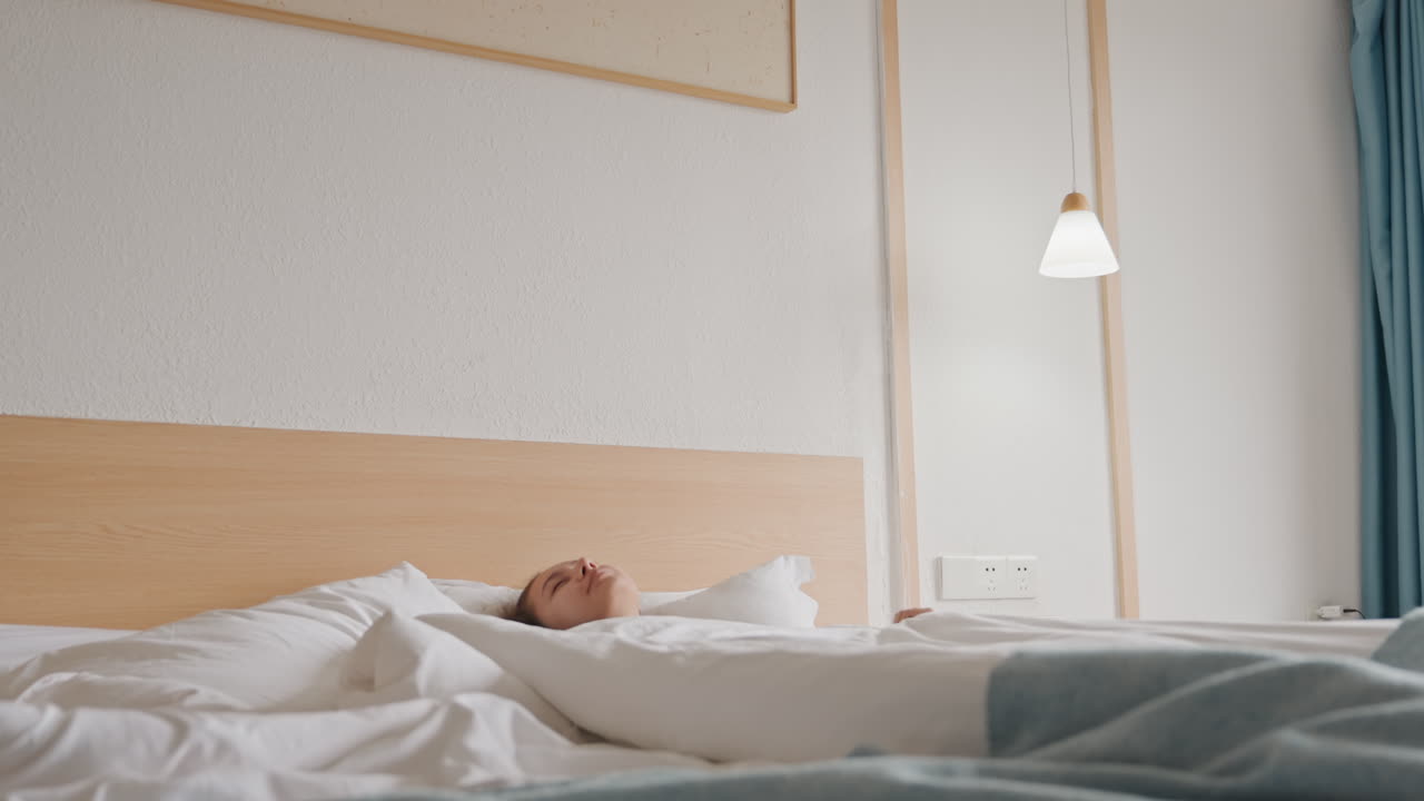 Donna assonnata nel letto d'albergo che si sveglia e poi si riaddormenta sotto un piumino bianco con lampada da comodino e tende chiare, si siede, espira, si accoccola nei cuscini, atmosfera tranquilla e minimalista della stanza.