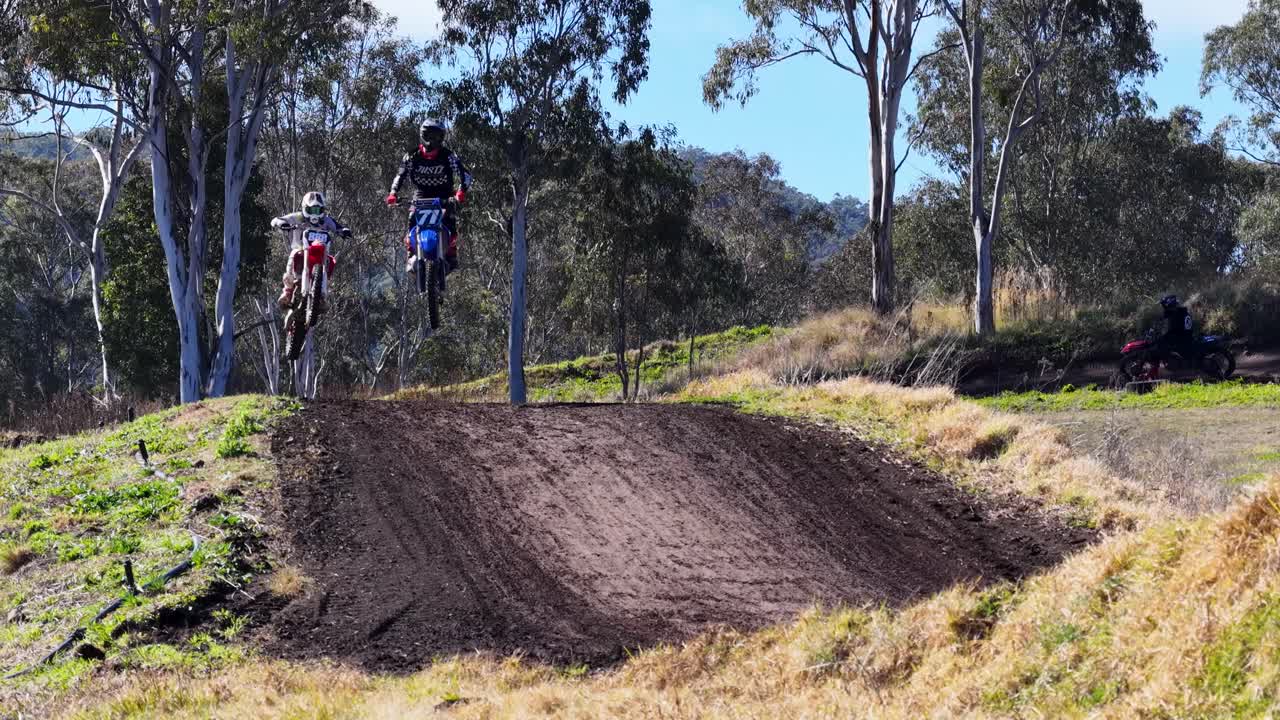 Motocross riders jump a dirt mound on a sunlit outdoor track, shown in wide shot