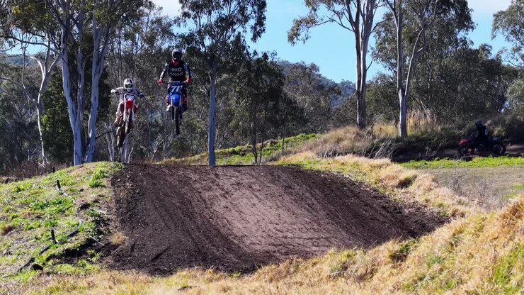 Motociclisti da motocross saltano su un cumulo di terra su una pista esterna illuminata dal sole, mostrato in campo lungo