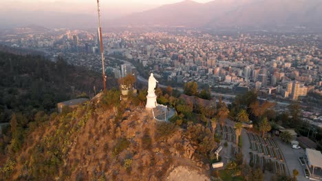 Aerial-pan-left-of-statue-in-Sanctuary-of-the-Immaculate-Conception-in-San-Cristobal-Hill-summit,-Santiago-city-in-background-at-sunset,-Chile