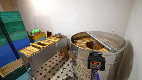 little room with honey extractor machine, metal box of frames and a pile of wooden hives. centrifuge working pumping honey. top view.