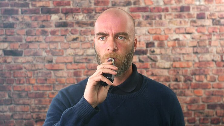 Ritratto di un uomo che svapa al chiuso davanti a un muro di mattoni, che guarda direttamente la fotocamera con una nuvola di fumo.