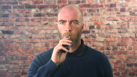 Retrato-De-Un-Hombre-Vapeando-Adentro-Frente-A-Una-Pared-De-Ladrillos-Mirando-Directamente-A-La-Cámara-Con-Una-Nube-De-Humo