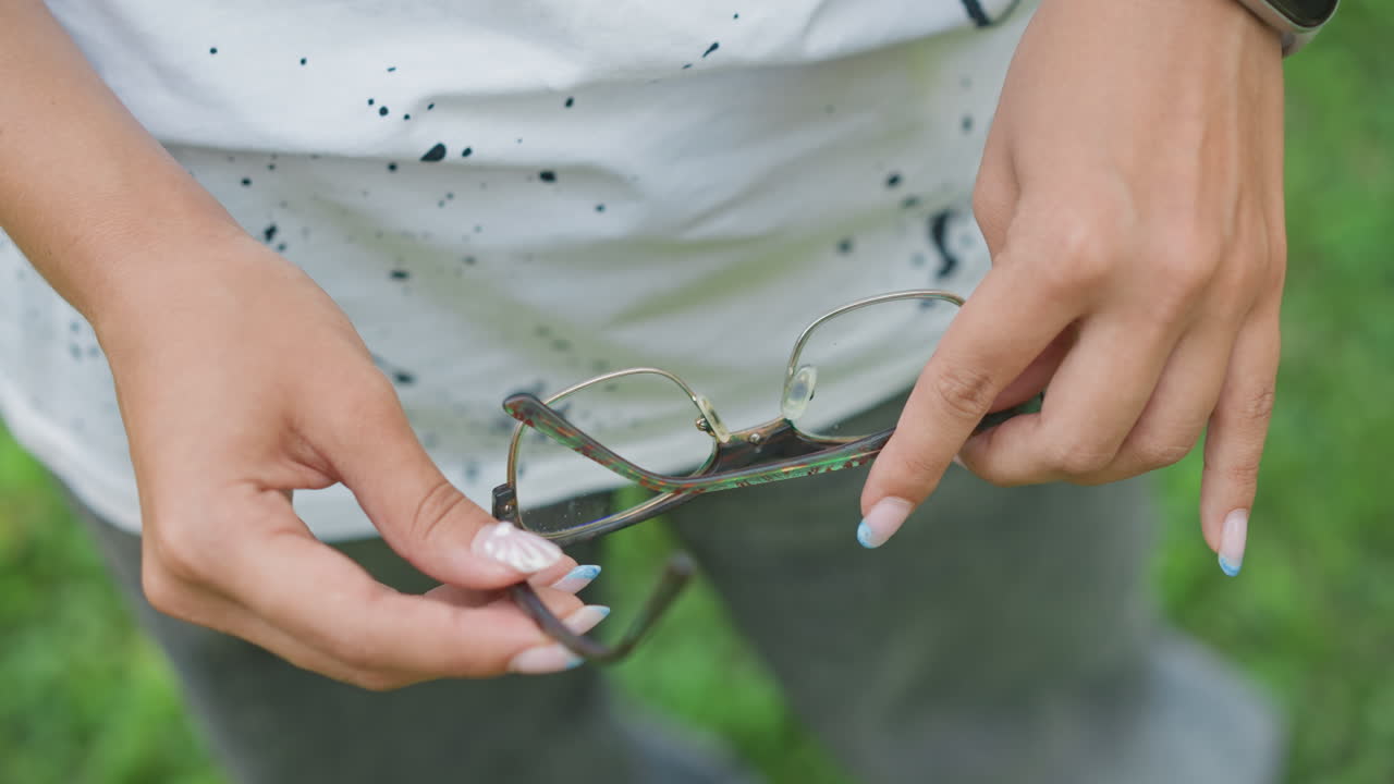 Mujer negra doblando cuidadosamente unas gafas de sol estampadas, parque con césped de fondo, manicura cuidada, cierre de bisagra meticuloso, accesorio listo para el bolsillo, hábito de viaje diario, momento diario de calma