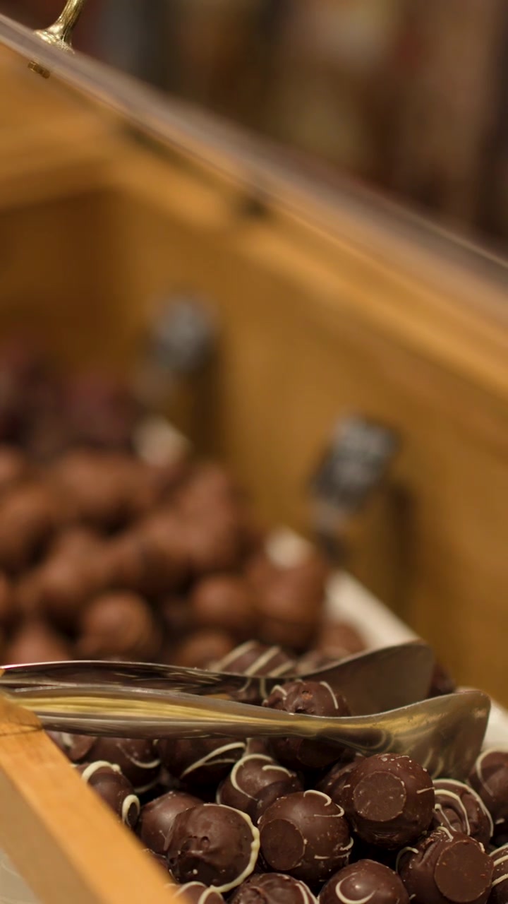 Hand choosing assorted chocolates from wooden display under warm lighting, shallow depth of field, Bruges
