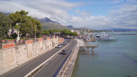 vista al mar del puerto deportivo costero de gaeta con barcos comerciales atracados en el puerto y tráfico de automóviles en la carretera frente al mar con la ciudad y las montañas en el fondo, italia, estática