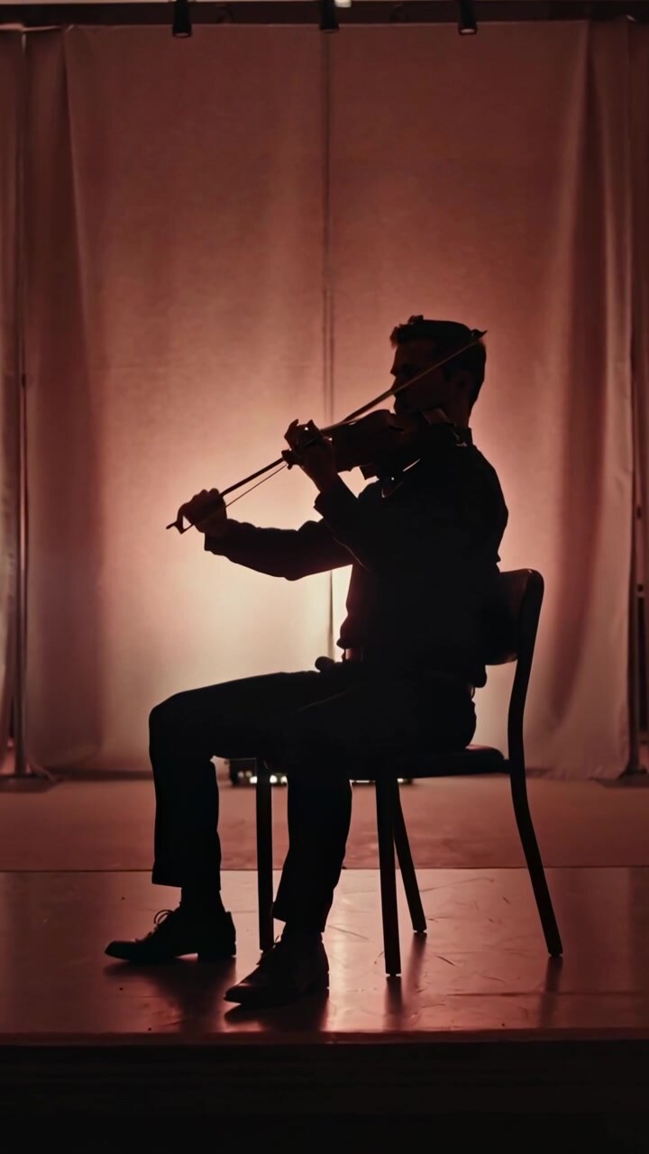 Video verticale: Violinista seduto che suona il violino con l'archetto sul palco, in abiti scuri formali.