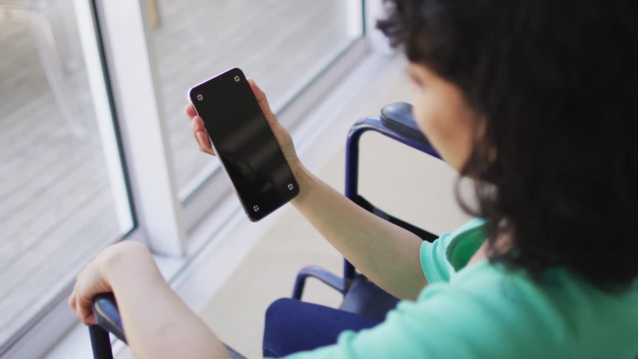 Biracial disabled woman in wheelchair using smartphone in living room, copy space on screen