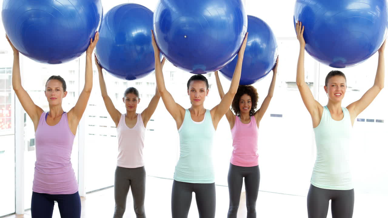 Fitness class in studio lifting exercise balls while squatting