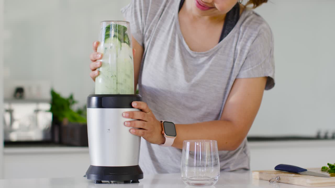 Donna che sistema la tazza, preme il frullatore, frullando cetriolo e verdure in una bevanda energizzante sul bancone.