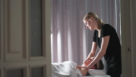 massage therapist in black uniform gently massaging chest of light-skinned female client lying under white sheet in serene spa room, soft natural light filtering through grey curtain at side of frame