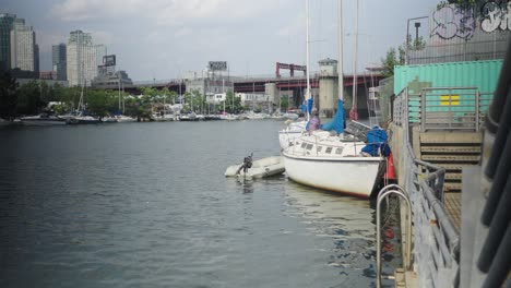 An-elderly-white-sailboat-with-a-single-hull-and-an-adjacent-parked-dinghy-rests-contentedly-against-the-canal-wall-in-Greenpoint,-Brooklyn