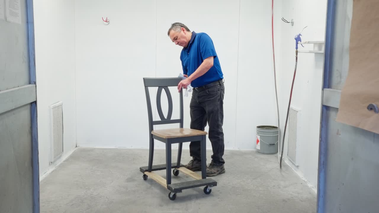 Static camera shows man sanding dining chair back in booth, prep for coating