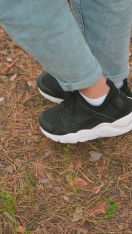 primer plano de dos individuos caminando por un sendero forestal con pantalones vaqueros azules, uno con zapatillas negras y el otro con zapatillas blancas, rodeados de agujas de pino, hojas y ramas pequeñas