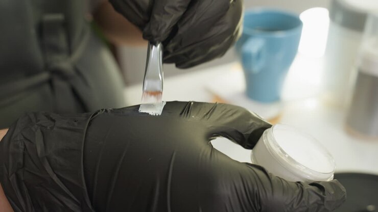 Closeup of beautician in black gloves dipping small angled brush into white cream jar and blending cream on glove in well-lit salon setting with tools and products blurred in background