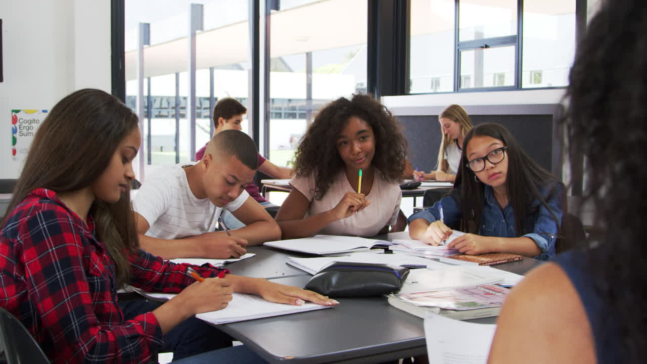 Alunos do ensino médio levantam as mãos, professor sentado à sua mesa