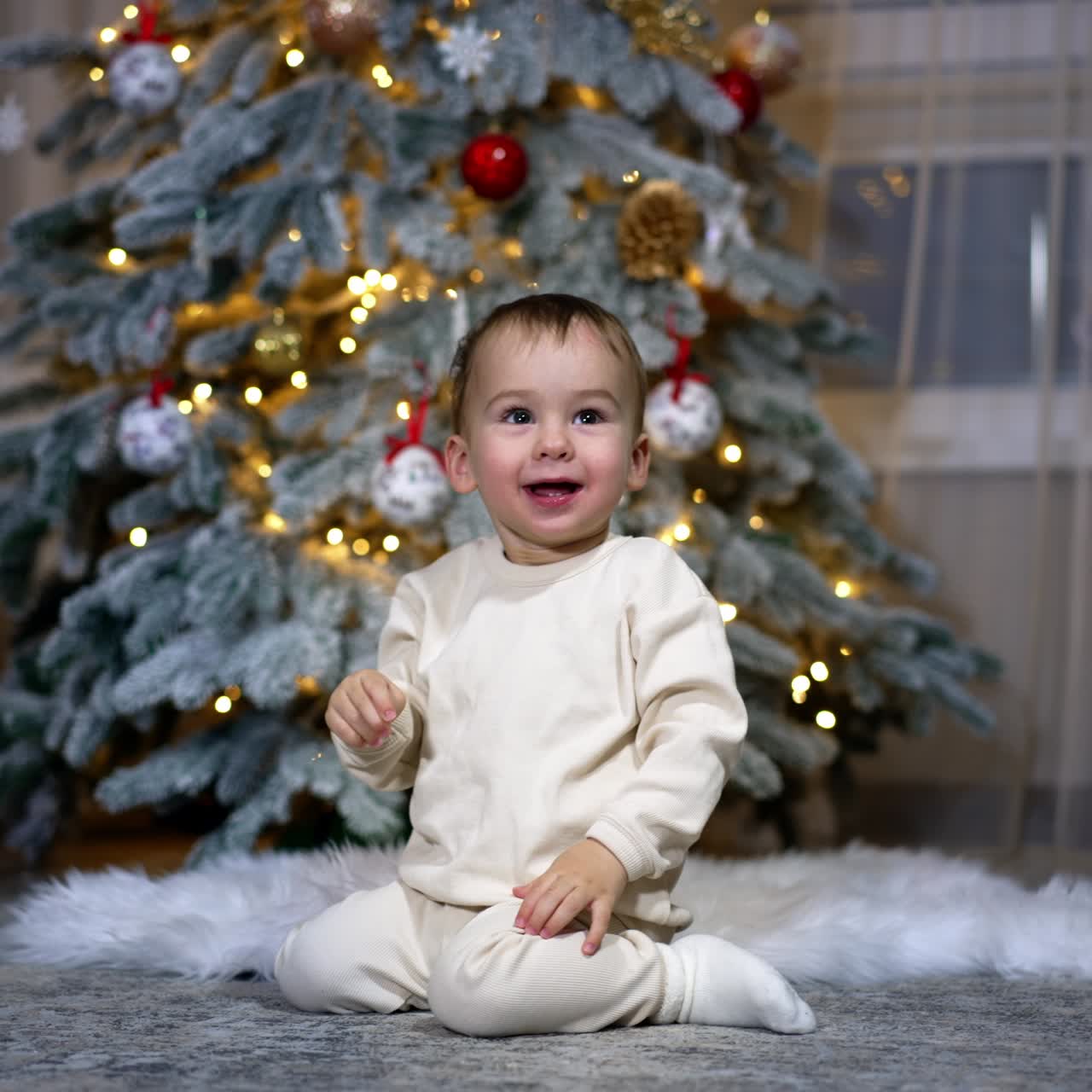 Happy beautiful excited kid waving his hands. Baby sits on the floor rejoicing and laughing adorably. Christmas tree at backdrop