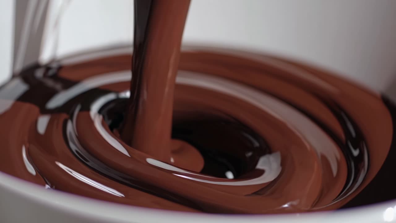 Video ravvicinato che riprende cioccolato fuso che si versa in una ciotola bianca, evidenziandone la consistenza liscia.
