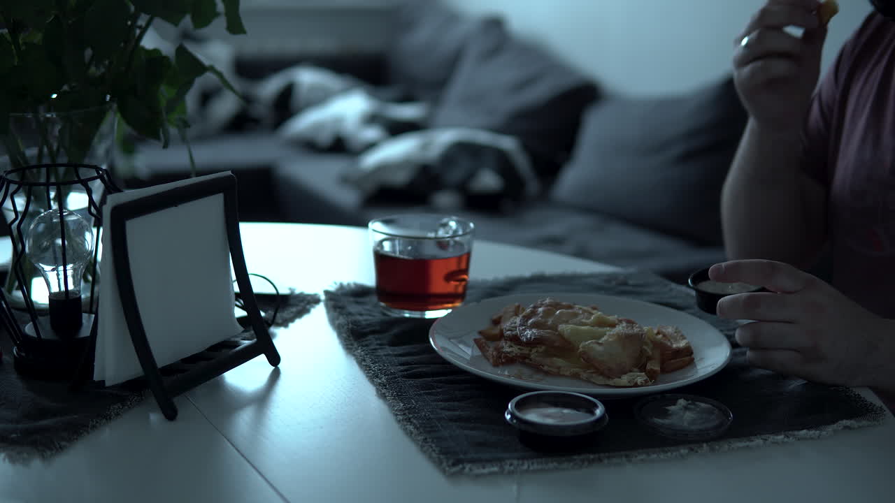 The man sits at the table and eats his meal, there is a white plate with fries and tea in a glass, the table is decorated with a vase with flowers and a napkin