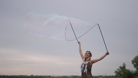 una joven hippie en un vestido y con plumas en la cabeza hace enormes burbujas de jabón al atardecer en la orilla de un lago en cámara lenta