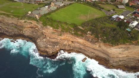 Toma-épica-De-Drones-De-Olas-Oceánicas-Rompiendo-Contra-Acantilados-Y-Rocas,-Cerca-De-Un-Parque,-Sydney,-Australia