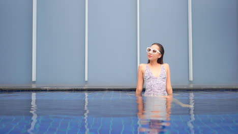 attractive asian woman wearing bathing suit and sunglasses sitting in clear shallow water of resort swimming pool on vacation