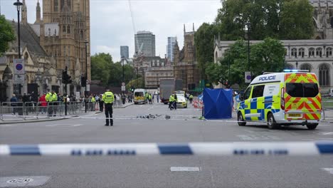 blue and white police cordon tape flutters in the wind in front of officers next to a van working the scene of a serious bicycle and truck collision incident in front of the houses of parliament.