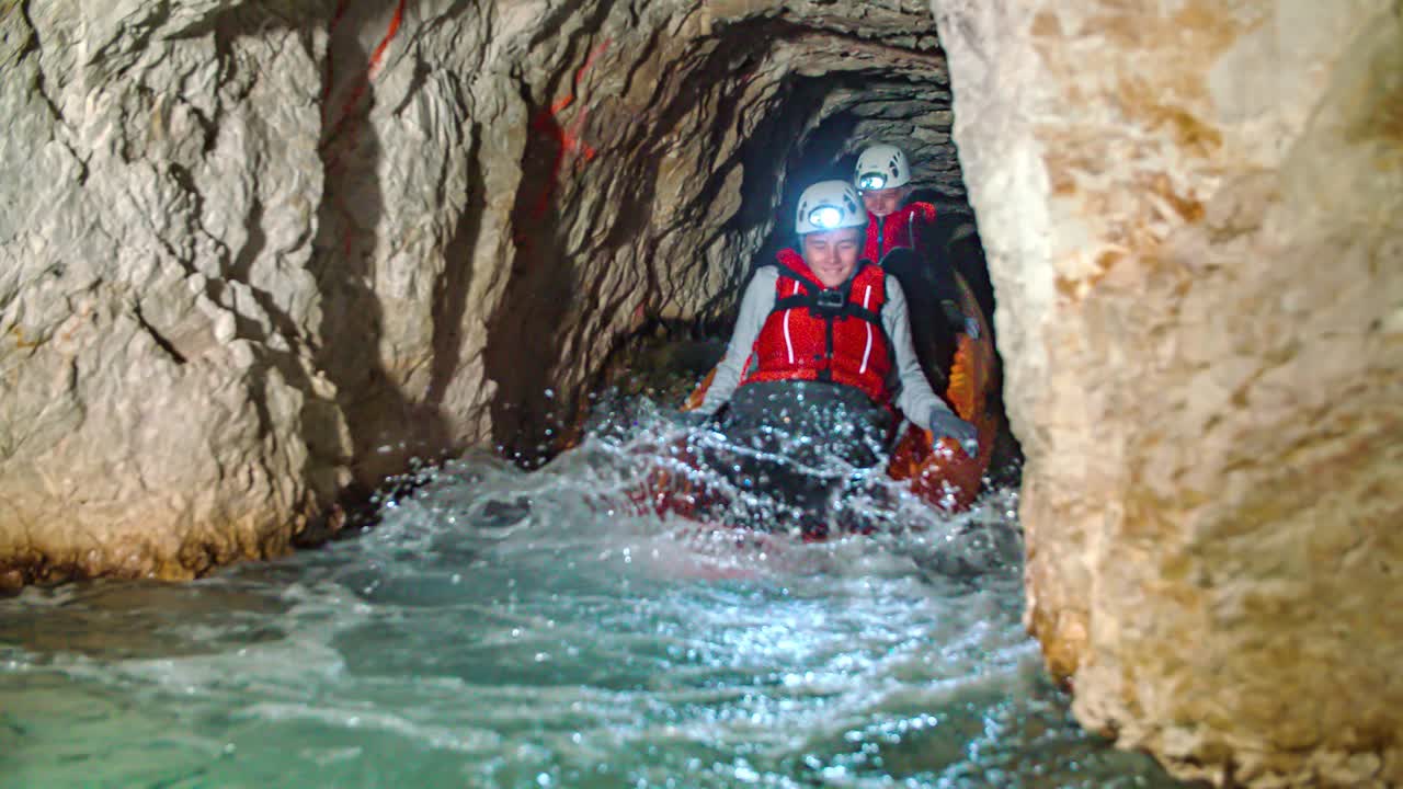 Two young cute Caucasian boys ride kayak down small narrow waterfall in underground rocky mining tunnel, Podzemlje Pece, Mezica, Slovenia, slow motion pan