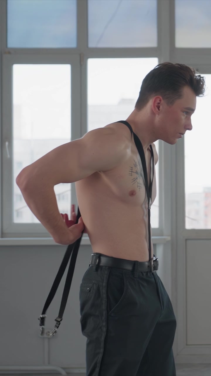 Giovane uomo a torso nudo sistema le bretelle nere sopra pantaloni scuri. L'uomo fissa le cinghie intorno alle spalle. Lo sfondo mostra una vista urbana attraverso vetri smerigliati e pareti grigie