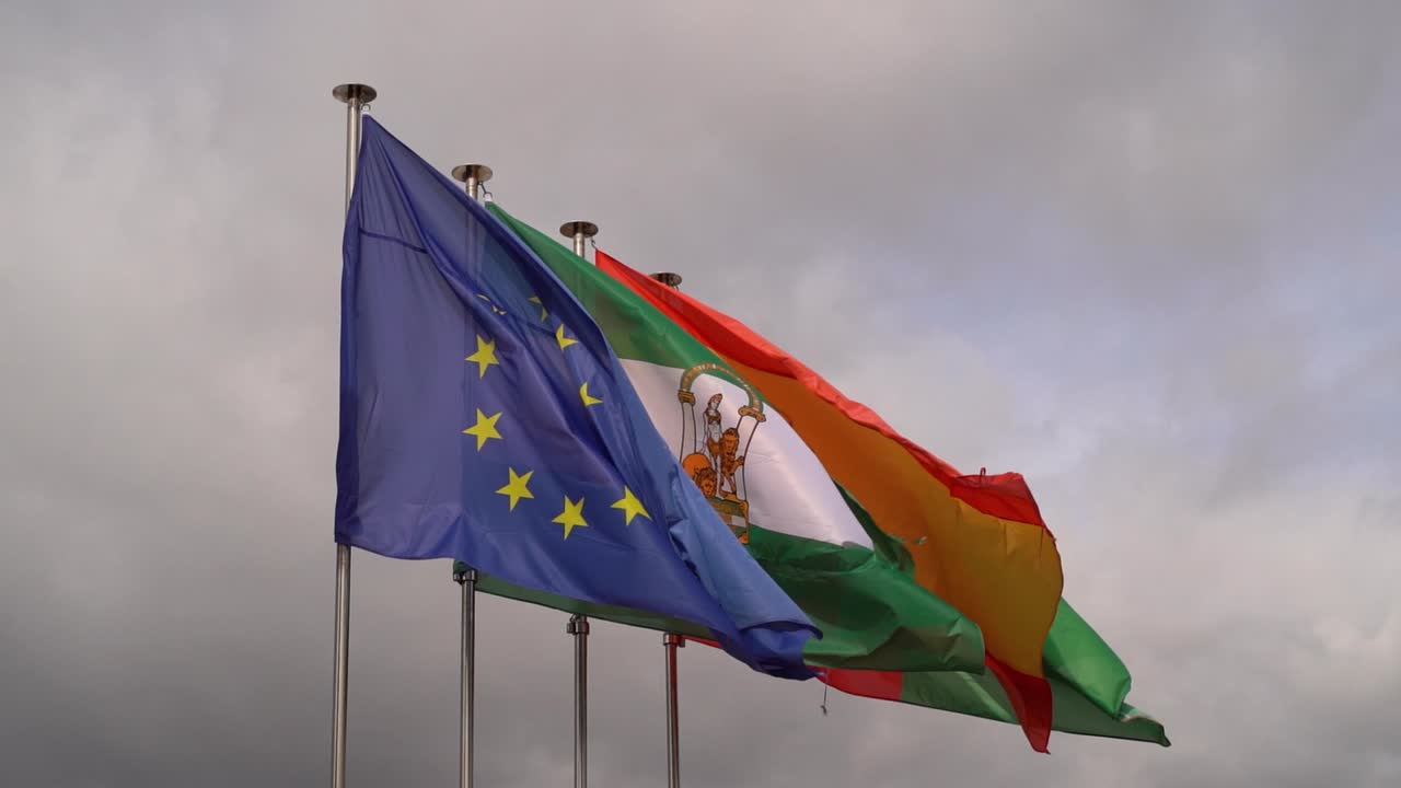 bandera europea frente a la bandera española y andaluza ondeando contra el cielo gris