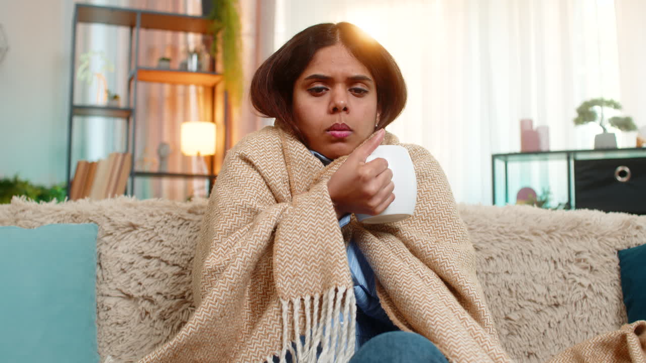 Indian woman drinking hot tea and coughing at home under blanket showing cold and tired expression